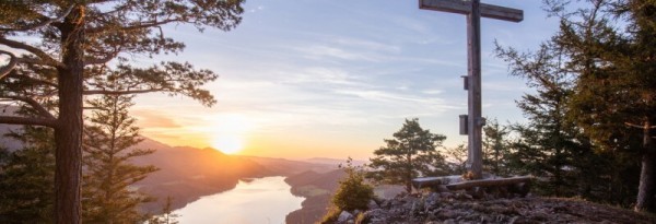 Holzkreuz auf einem Berggipfel mit Blick auf den Fuschlsee und Berge bei Sonnenuntergang, umgeben von Bäumen.
