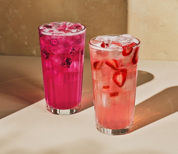 Two iced drinks in tall glasses: one dark pink with floating dragonfruit pieces, the other light pink with floating strawberry pieces.