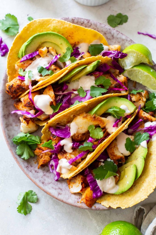 Four fish tacos with red cabbage, avocado, cilantro and fish taco sauce on a plate.