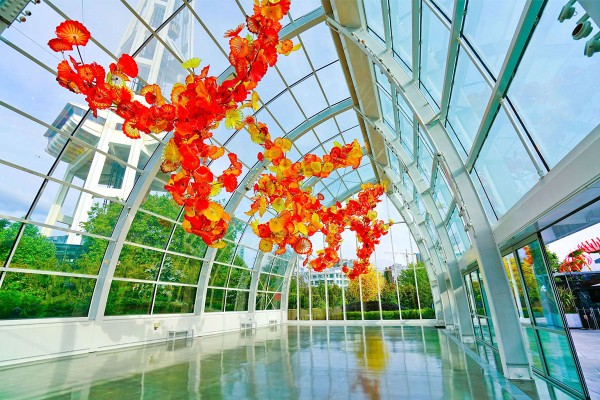 A spacious glass atrium with vibrant orange and yellow glass sculptures suspended from the ceiling. The transparent walls reveal a green garden outside.