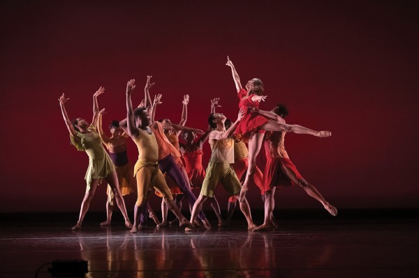 Dancers in colorful outfits perform a dynamic routine on stage. One dancer in red leaps gracefully in the center, while others in yellow, green, and orange strike expressive poses against a deep red backdrop.