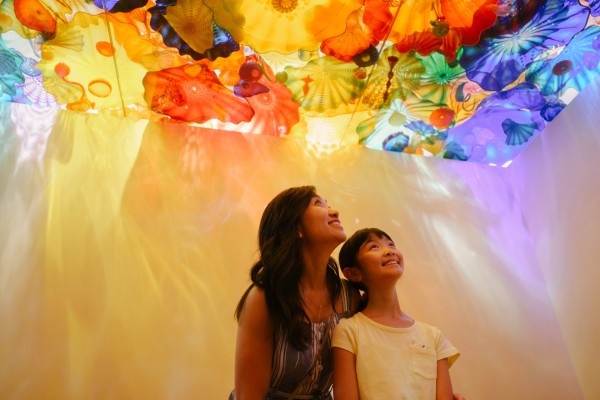 A woman and a young girl gaze up, smiling, at a colorful, translucent ceiling installation resembling glass flowers. The room is bathed in warm, vibrant hues of orange, yellow, blue, and purple.
