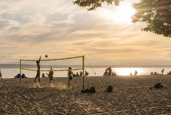 People playing beach volleyball under a partly cloudy sky during sunset. The golden light reflects on the water, creating a warm and serene atmosphere. Other individuals are relaxing on the sand in the background.