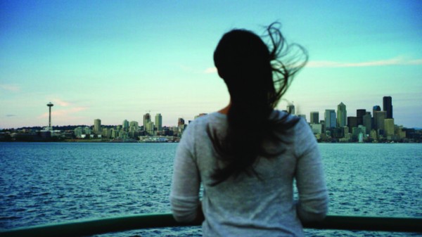 A person with long hair stands at a railing, facing a large body of water with a city skyline in the background. The Space Needle is visible to the left. The sky is clear, and the wind is blowing their hair.