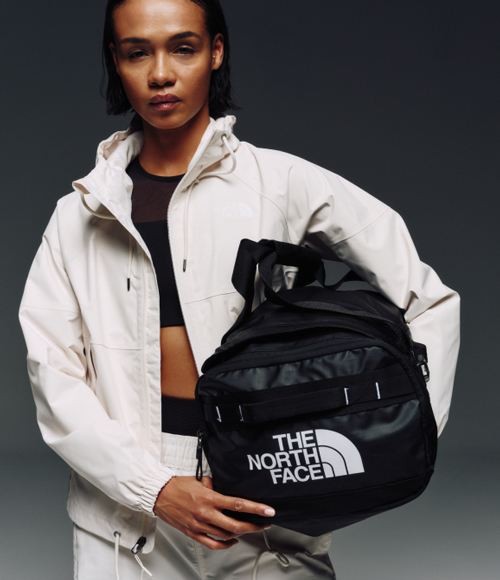  A woman in a white jacket and a black crop top holding a black duffel bag and looking at the camera. 