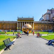 在晴朗的蓝天下，游客可以在院子里的长椅上放松身心，院子里有一座大型青铜雕塑和历史悠久的建筑。