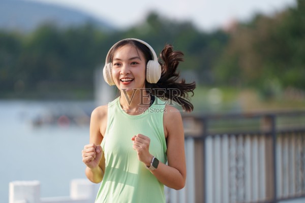 一位年轻的亚洲运动女性戴着耳机，沿着宁静的湖边慢跑，享受着音乐，她身穿浅绿色背心，佩戴着智能手表，脸上洋溢着快乐与活力。照片摄影图片
