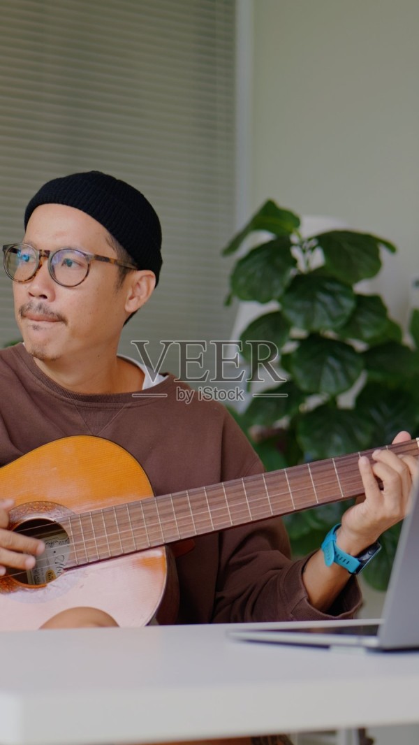 亚洲男性在现代办公桌上边使用笔记本电脑边快乐地弹吉他，健康<a target=