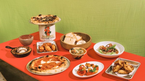 A spread of Italian-inspired dishes arranged on a red table with a green textured backdrop. The assortment includes two pizzas, one topped with pepperoni and another placed on a can of Bianco DiNapoli tomatoes, a skillet cookie with ice cream, tiramisu in a striped bowl, crispy chicken wings on metal trays, a wooden bowl filled with bread slices, a plate of tomato and burrata salad, a bowl of meatballs, and a dish featuring stuffed peppers with melon slices.